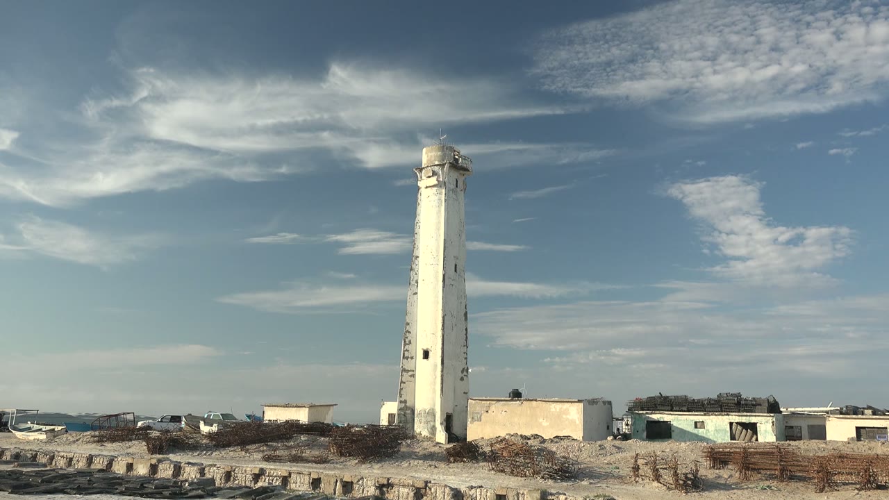 The Guerrero Negro lighthouse remains near the center of the frame beside low pale buildings, rough sandy ground, and broken clouds, with no visible people.