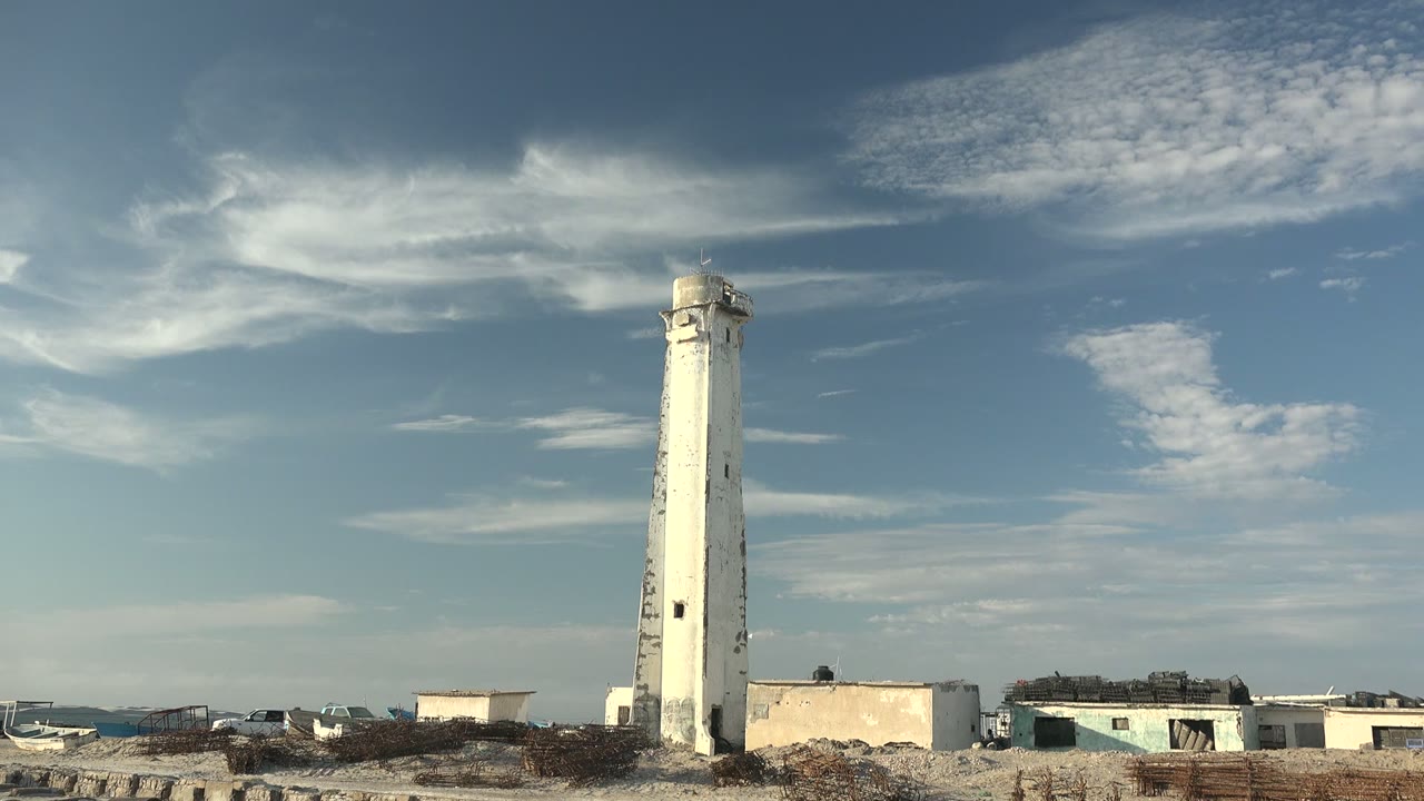 A tall white lighthouse in Guerrero Negro rises above flat sandy shoreline, small buildings, brush fencing, and a high blue sky with streaked clouds, with no visible people.