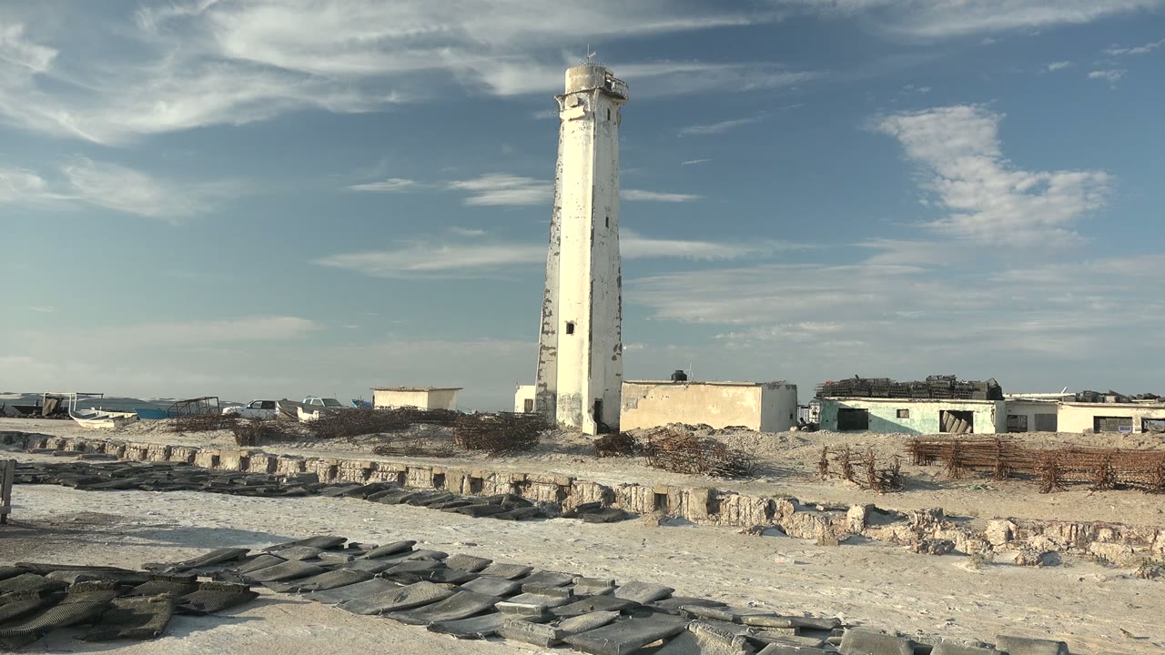 At 0:00, a white lighthouse stands on a sandy rock-strewn beach in Guerrero Negro, Baja California Sur, with small buildings behind it and no visible people.