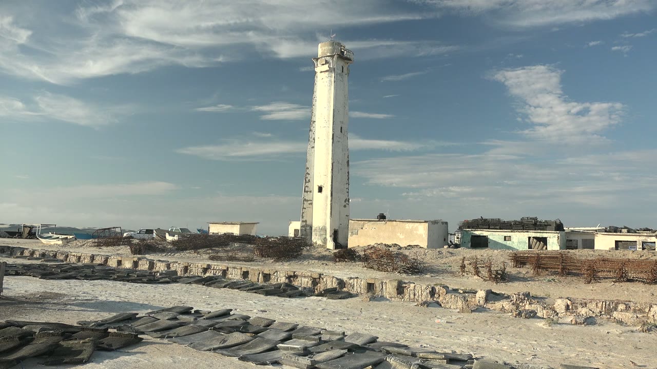 At 0:02, a tall white coastal lighthouse rises above sandy ground, rough stone formations, and low background buildings in Guerrero Negro, with no visible people.