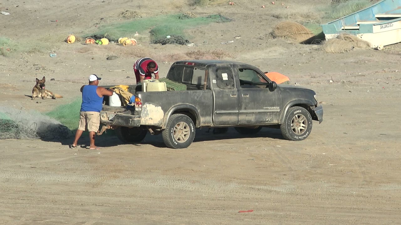 At 0:02 in San Juanico, Baja California Sur, a man at the tailgate reaches toward gear while another bends over the truck bed and the dog lies farther back.