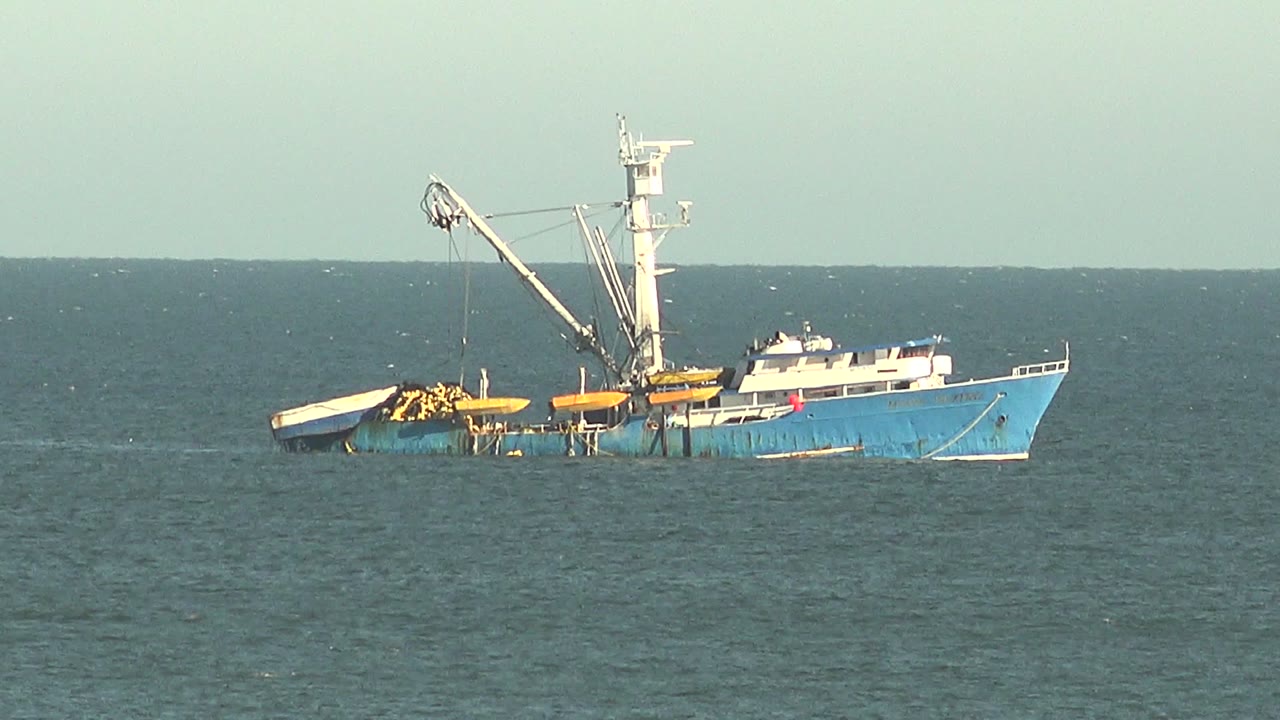 A blue work boat with a white cabin and crane sits under a pale sky off San Juanico, with small white wave flecks around it and no visible people.