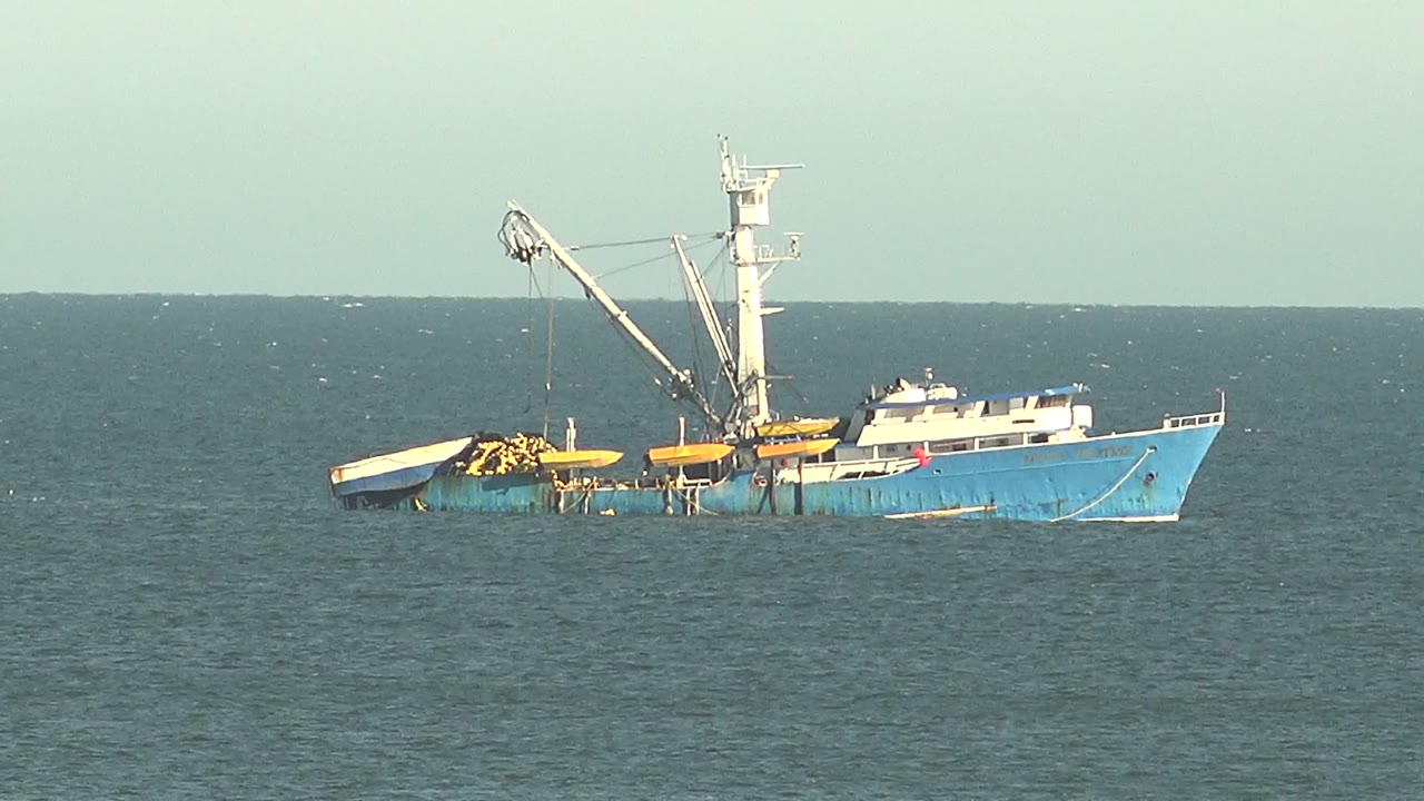 A blue fishing vessel sits partly submerged off San Juanico, Baja California Sur, with a crane raised over yellow deck structures and no visible people.
