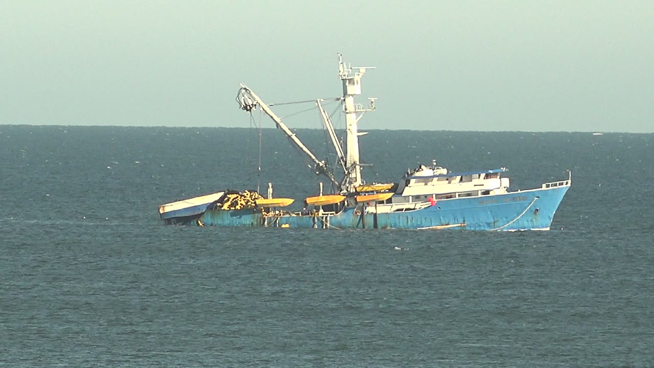 A partly submerged blue fishing boat holds in the midground off San Juanico, Baja California Sur, with the crane angled above the deck and no visible people.