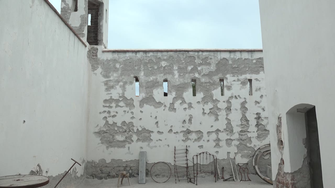 Mulege courtyard view with peeling white walls, narrow wall openings, a crenellated tower, tools and a wheel near the ground, and no visible people.