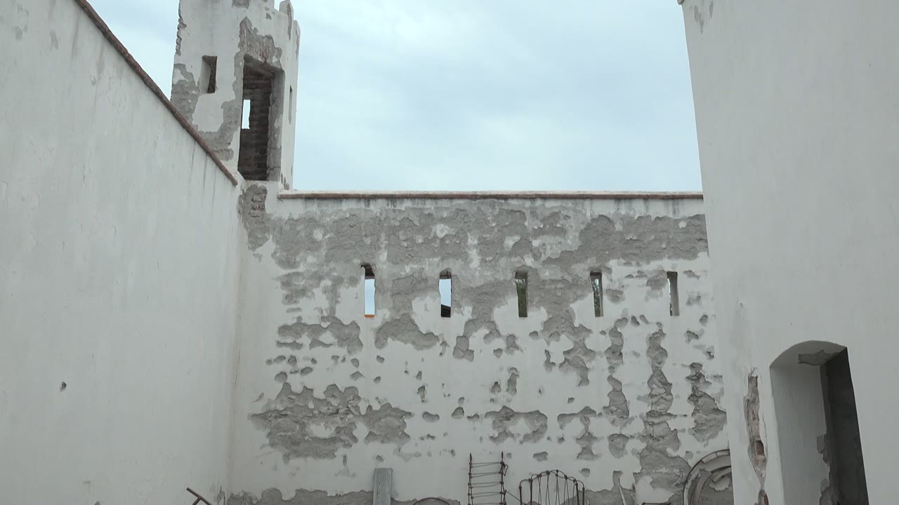 Mulege courtyard view showing the crenellated tower, mottled gray plaster patches, narrow openings in the wall, a cropped doorway at right, and no visible people.