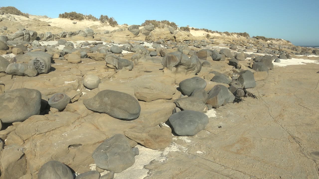 At La Bocana, Baja California Sur, smooth gray boulders sit across pale rock and sand under clear blue sky, with no visible people.
