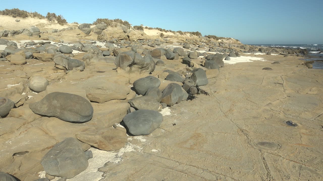 At La Bocana, Baja California Sur, flat pale rock leads toward clustered boulders and a broader strip of ocean waves on the right, with no visible people.