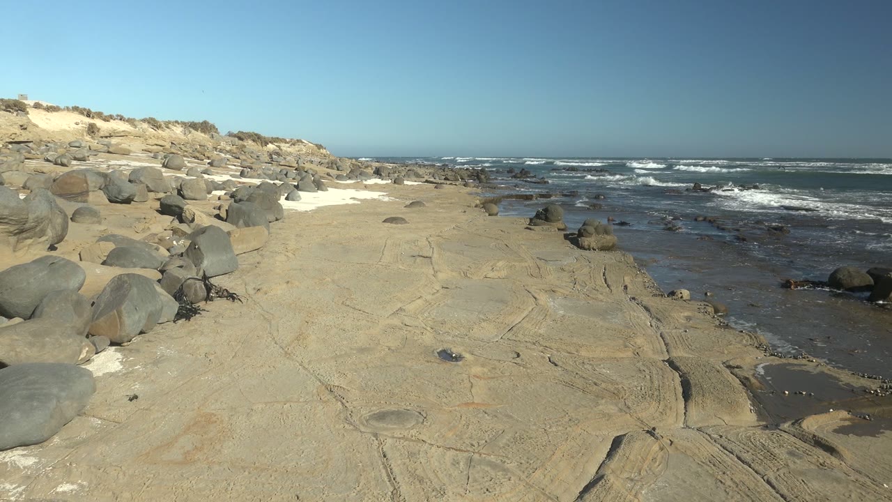 Pale rocky shore at La Bocana, Baja California Sur, with rounded boulders on the left, dark wet tide pools near the surf, white waves offshore, and no visible people.