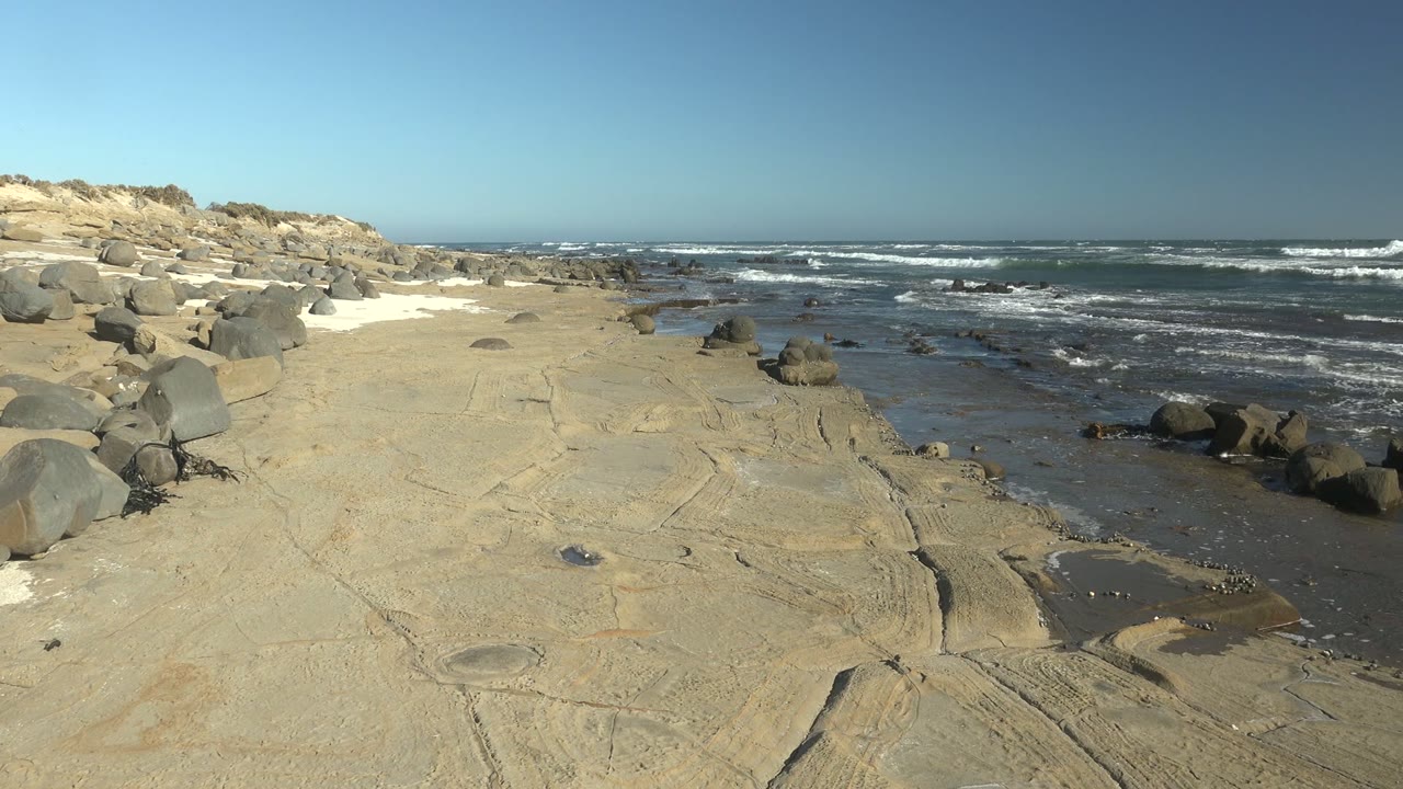 The La Bocana coast appears slightly farther along the same shoreline, with pale rock, left-side boulders, dark wet sand, and multiple white surf bands, with no visible people.