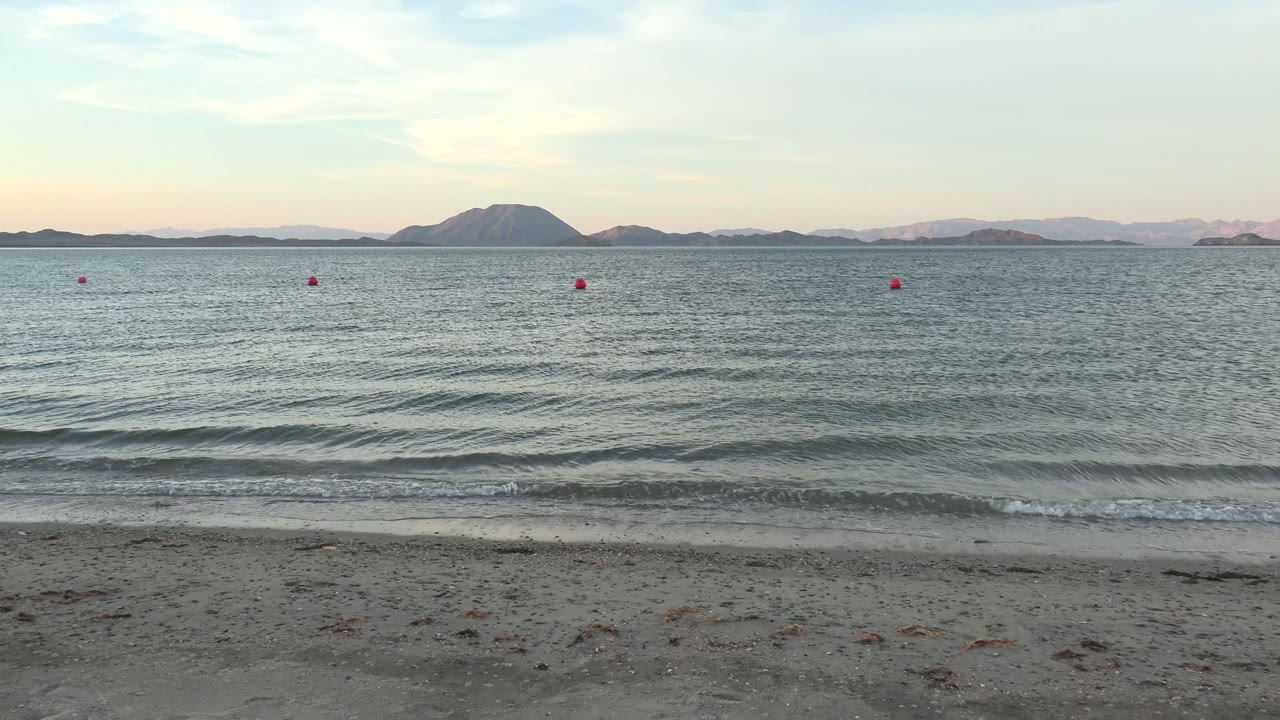 Wide coastal view in Bahia De Los Angeles with rippled water, a narrow band of foam at the beach edge, multiple red buoys, low island shapes, and no visible people.