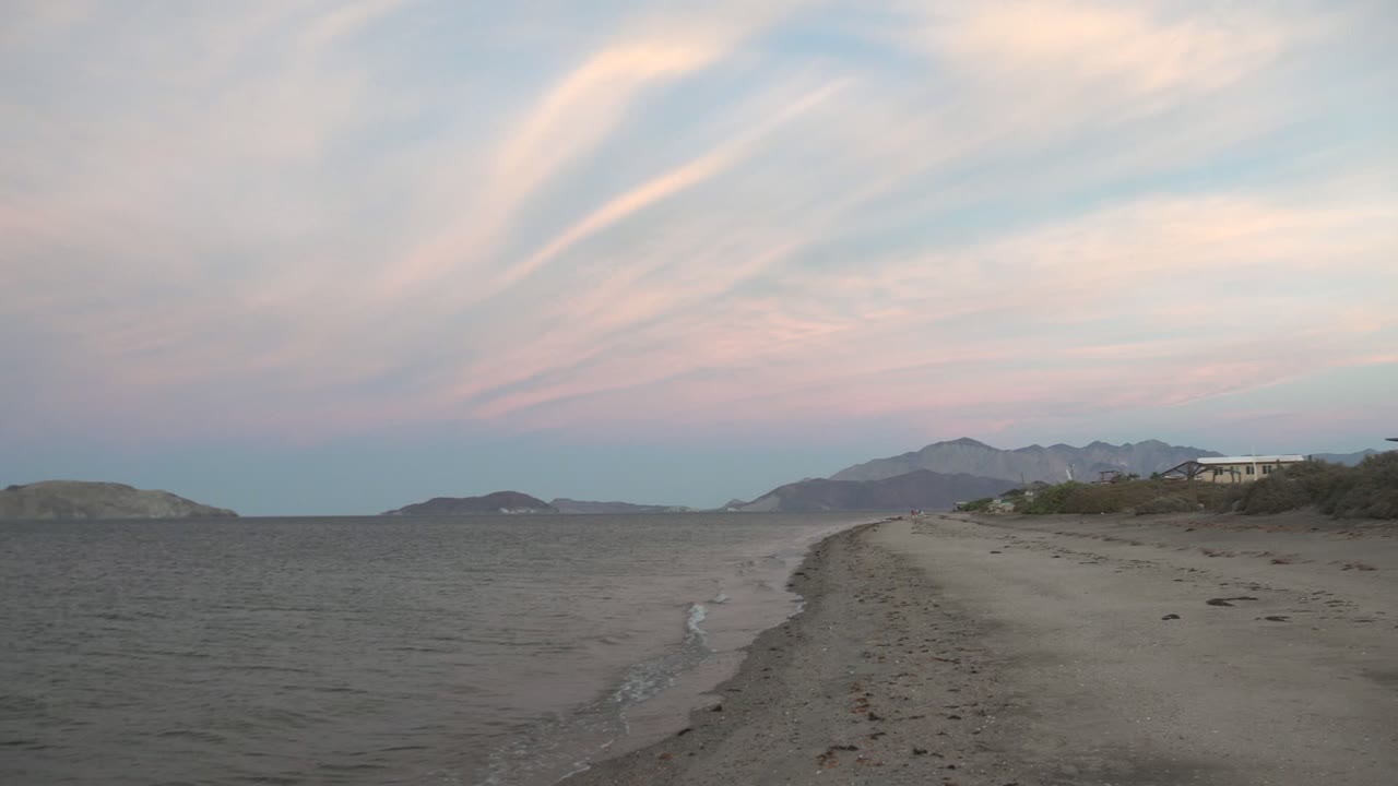 Bahia De Los Angeles sandy beach curves beside dark rippled water under pink-blue dusk clouds, with distant hills and no visible people.