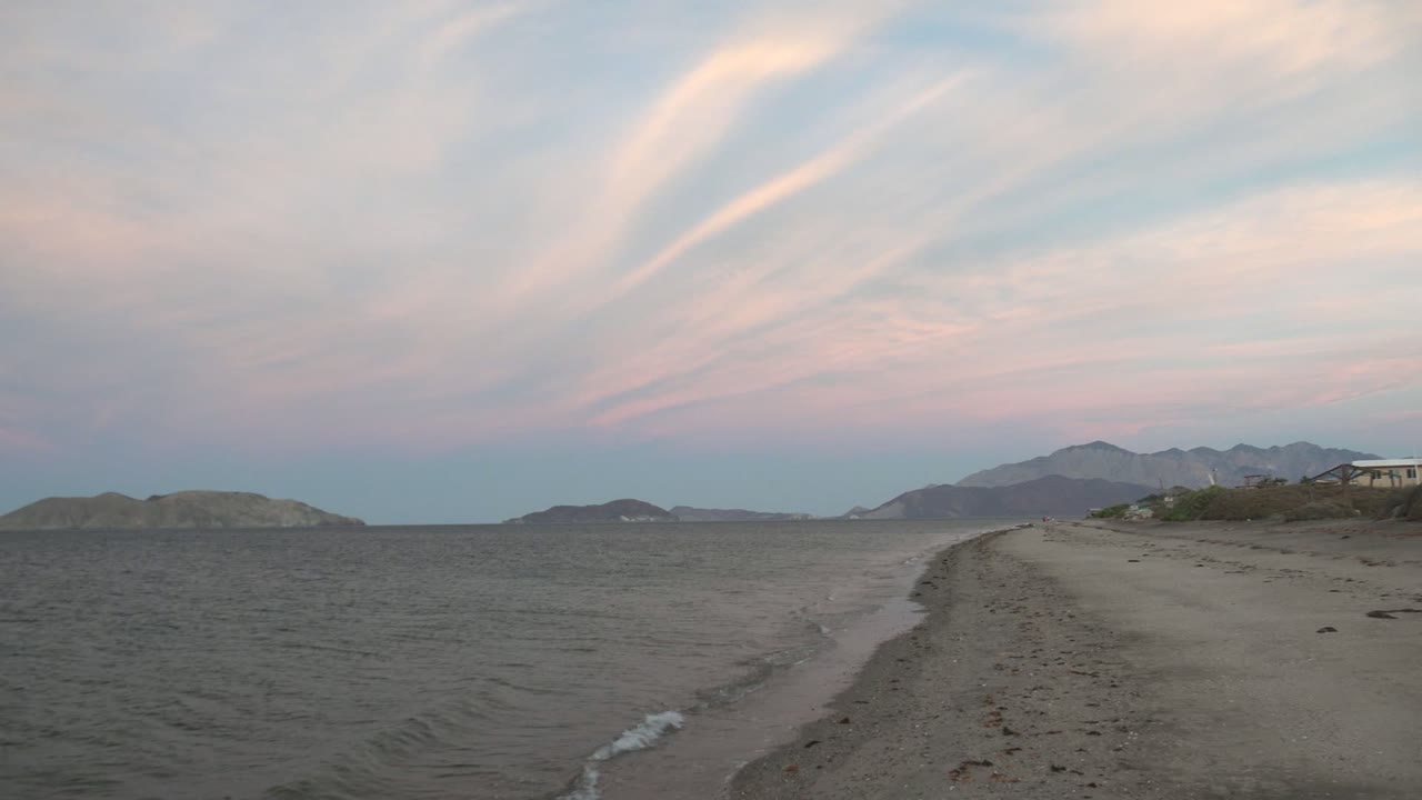 Bahia De Los Angeles beach sits beside rippled water as streaked pink clouds stretch over low hills and mountains, with no visible people.