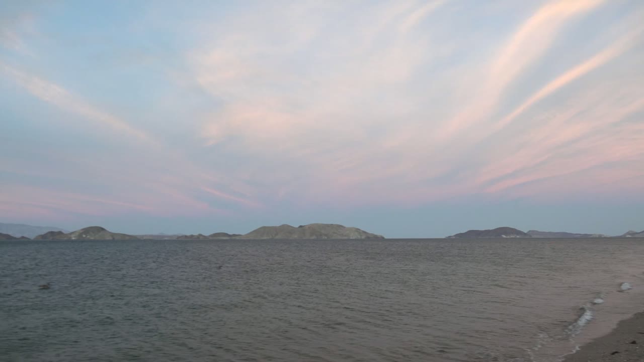 Bahia De Los Angeles ocean view with rippled water, distant island silhouettes, pink-blue clouds, a small sandy beach edge at lower left, and no visible people.