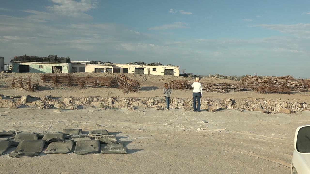 At 0:00 in Guerrero Negro BCS Mexico, a child and an adult stand in the distance across sandy ground with rubble, low damaged buildings, and a pale vehicle edge in the foreground.