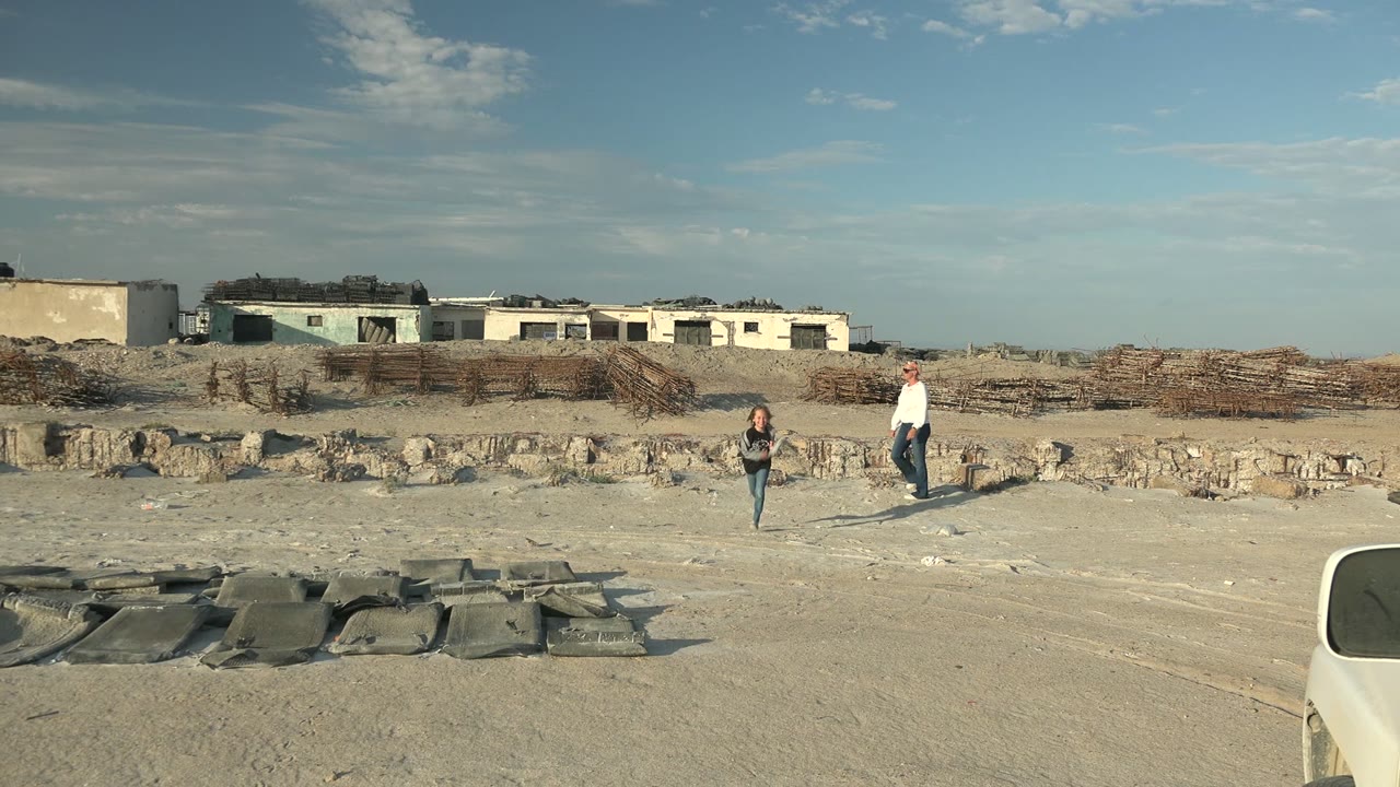 At 0:02 in Guerrero Negro BCS Mexico, the child and adult walk closer to the camera across the open sandy site, with stacked brush or debris and low buildings along the horizon.