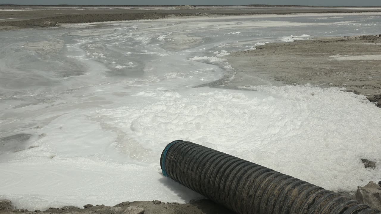 At 0:00 in Guerrero Negro, a ribbed black pipe points into foamy white water beside gray lagoon flats, with muddy ground at the edge and no visible people.