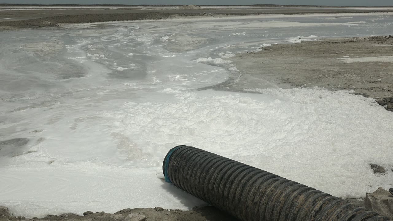 At 0:02 in Guerrero Negro, the dark corrugated pipe cuts diagonally through the lower frame as foamy discharge fills the center and gray mud flats sit beyond, with no visible people.