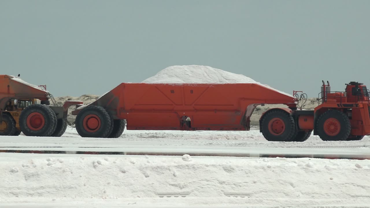 Red haul trucks in Guerrero Negro, Baja California Sur move side-on across a white salt flat with piled white material and no visible people.