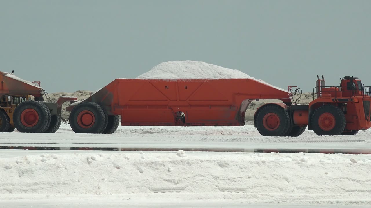 A red industrial tractor sits beside a red haul trailer on the white Guerrero Negro flat, with low earth berms in the background and no visible people.