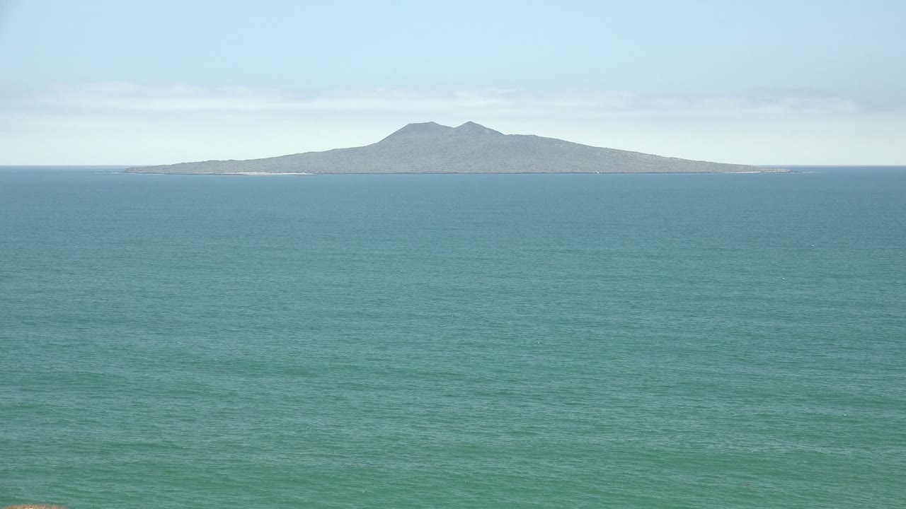San Quintin BC Mexico frame with the island held near the center of the horizon, broad rippled ocean water below, thin cloud cover overhead, and no visible people.