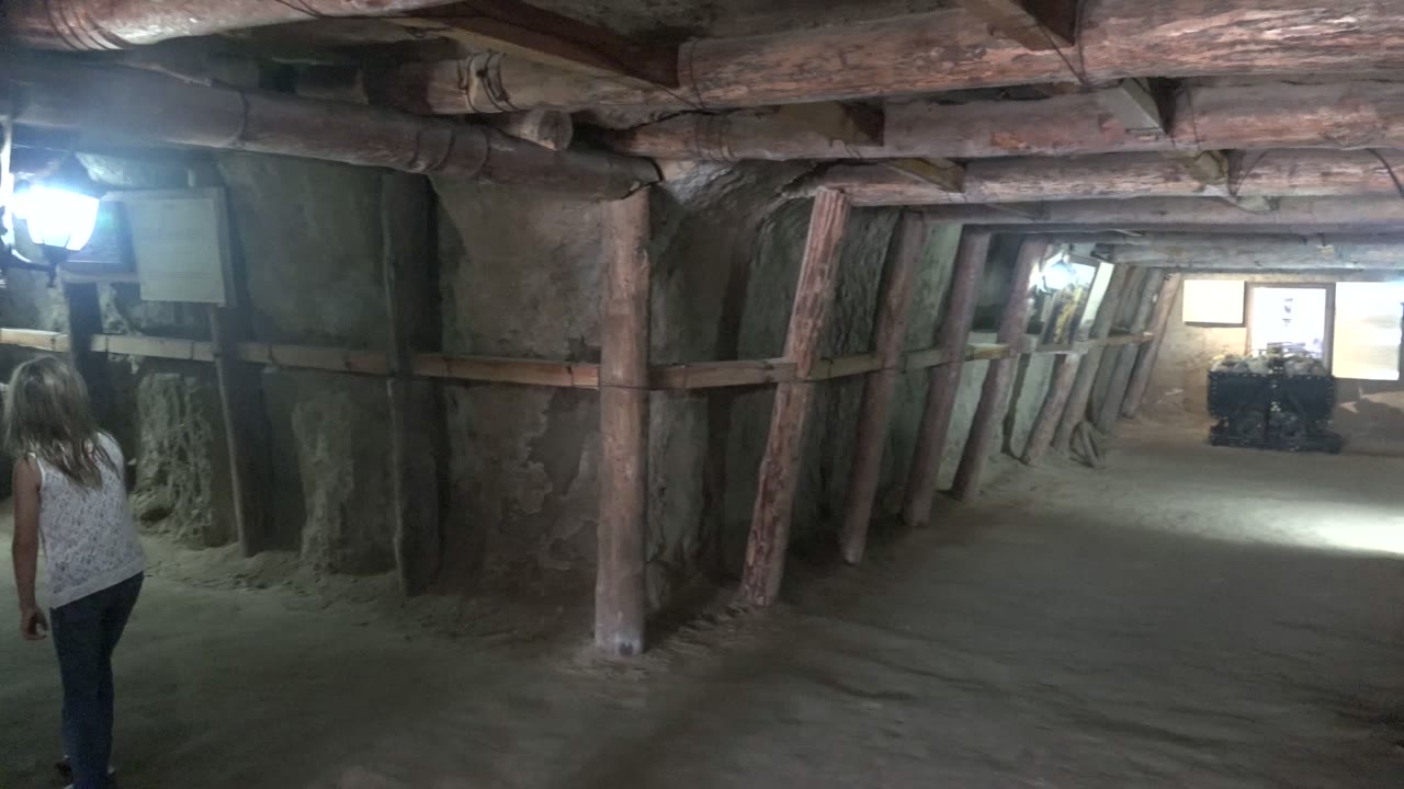 A person in a white top walks away at the left edge of a dim underground tunnel in Santa Rosalia, Baja California Sur, with timber posts, rough stone walls, and a bright lamp ahead.