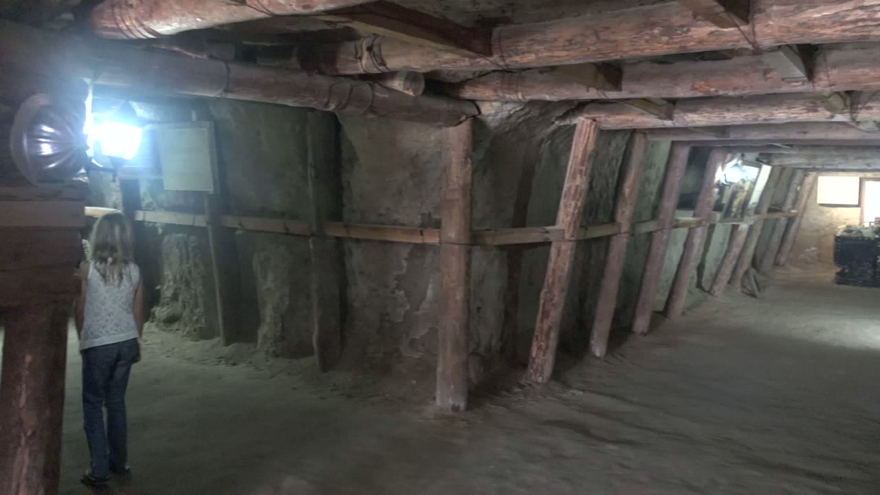 Person farther down a dim tunnel with wooden supports and bright light.