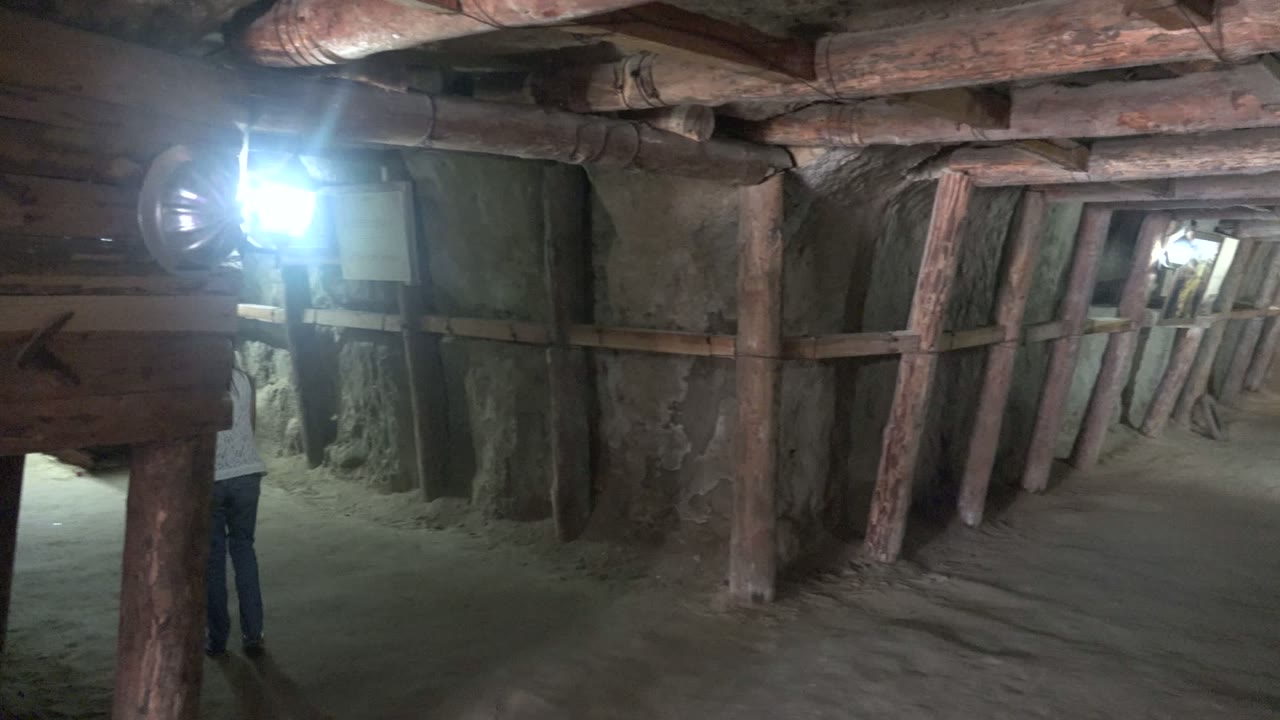The person is partly obscured near the right side of the Santa Rosalia tunnel, while a large wooden structure, vertical posts, rough stone surfaces, and a bright left lamp dominate the frame.