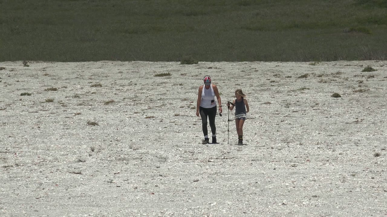An adult and child appear small in a wide view of a pale rocky plain in San Carlos, Mexico, with a dark flat background band behind them.