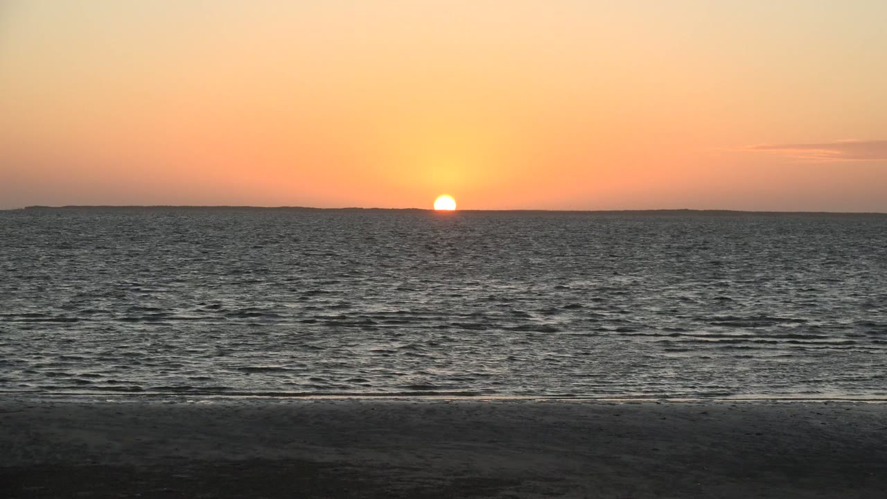 At 0:00 in San Carlos, Baja California Sur, the orange sun sits just above a low horizon across rippled ocean water with dark sand in the foreground and no visible people.