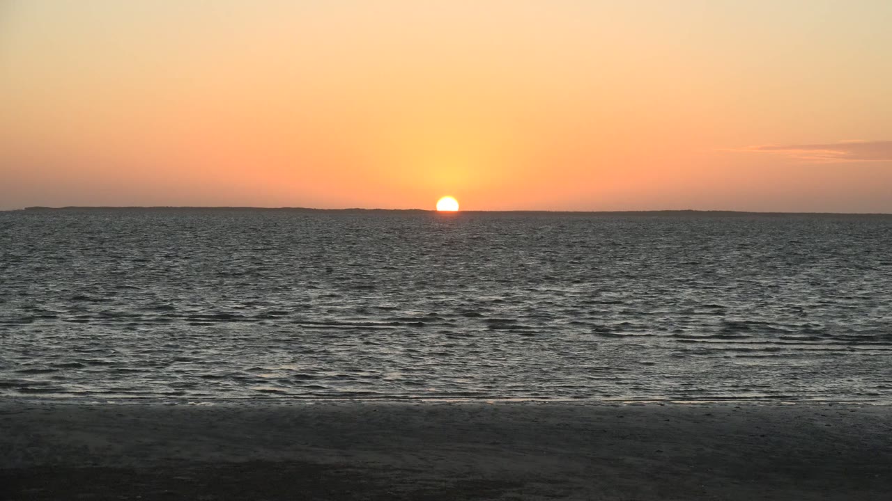 At 0:02 in San Carlos, Baja California Sur, the sun touches the distant horizon while darker ripples spread across the sea and the foreground beach stays in shadow, with no visible people.