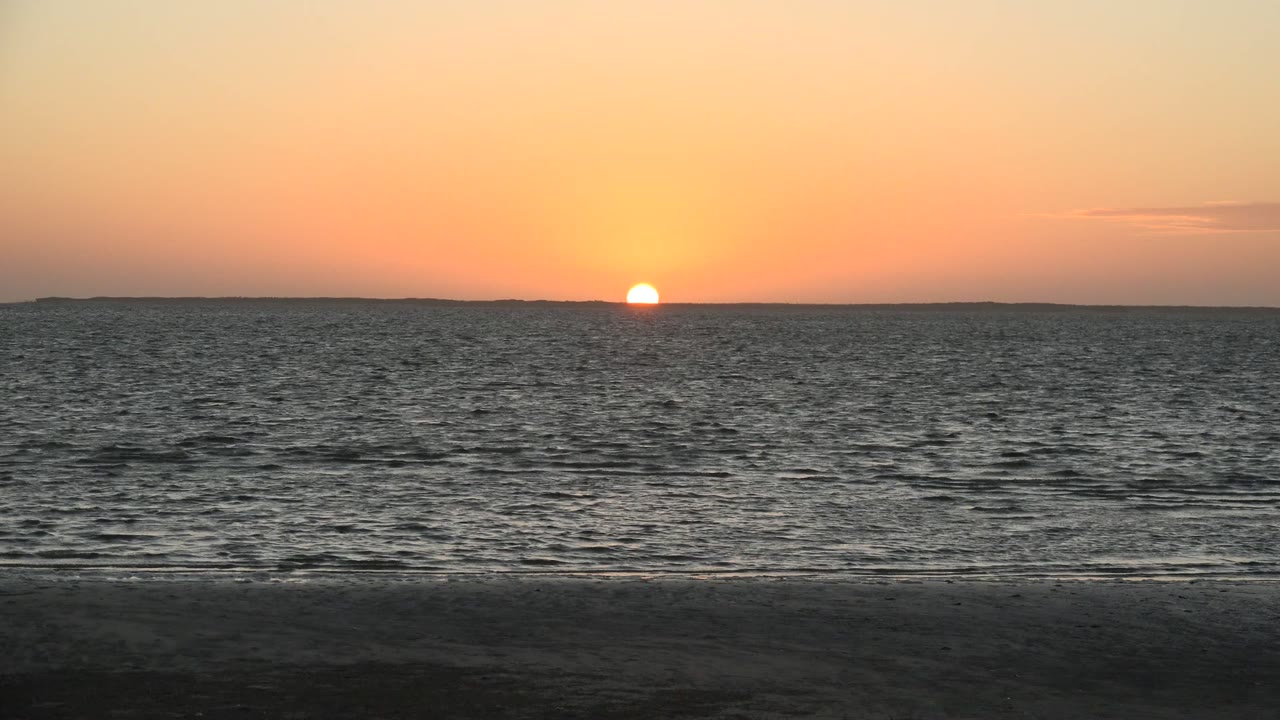 Low orange sun sits near the San Carlos, Baja California Sur horizon above rippled ocean water and a dark sandy beach, with no visible people.