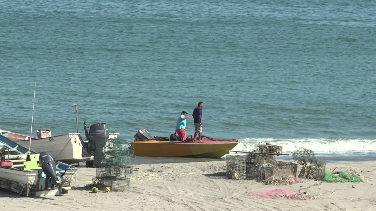 Two people stand and bend on a small boat at the San Juanico beach shoreline, with traps, nets, sand, and blue ocean water behind them.