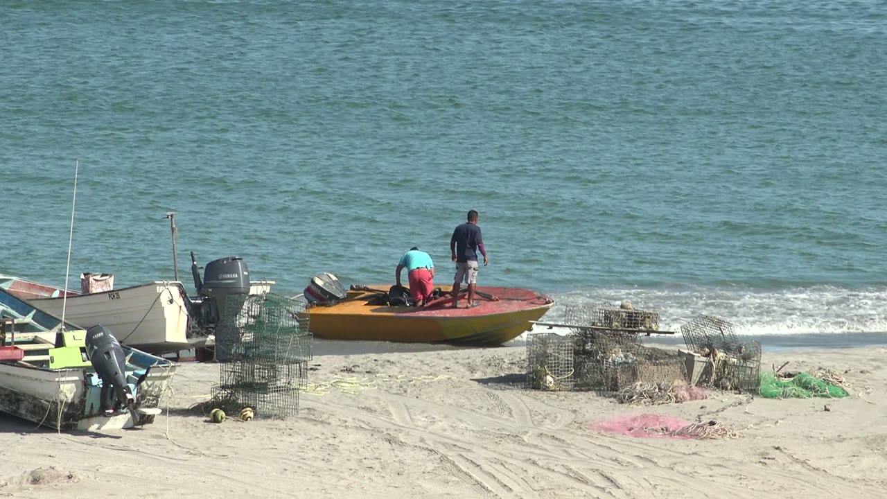 The small fishing boat remains at the San Juanico water's edge as one person bends low on the deck and the standing person faces the ocean.