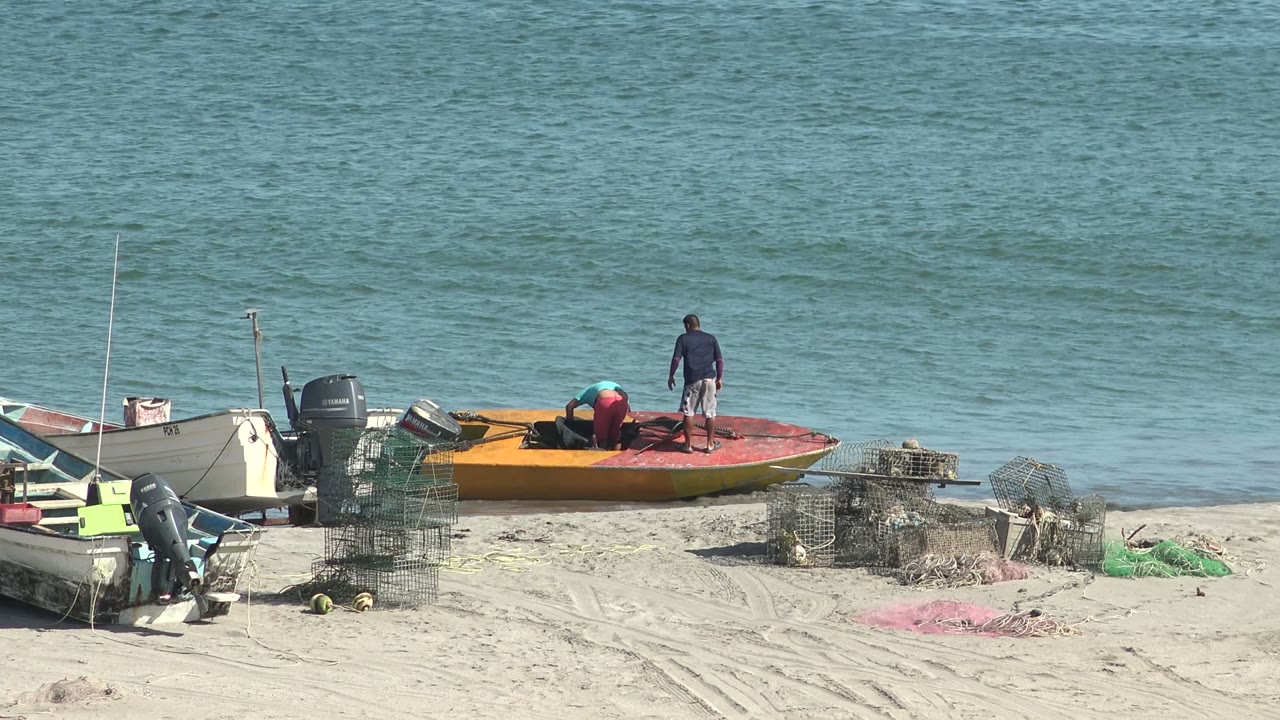 Two people work around an orange boat on the sandy shore in San Juanico, Baja California Sur, with crab traps, nets, other small boats, and calm ocean behind them.
