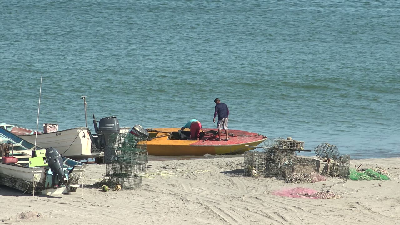 One person bends over gear on the orange boat while another stands near the stern, with small boats and stacked crab traps on the San Juanico beach.