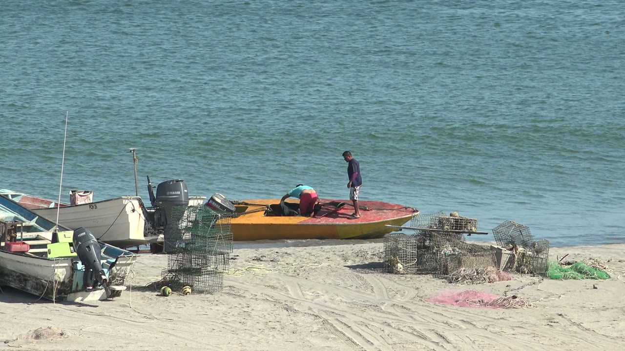 The two people continue working on the orange boat near the waterline in San Juanico, with the ocean surface behind them and fishing traps scattered on the sand.