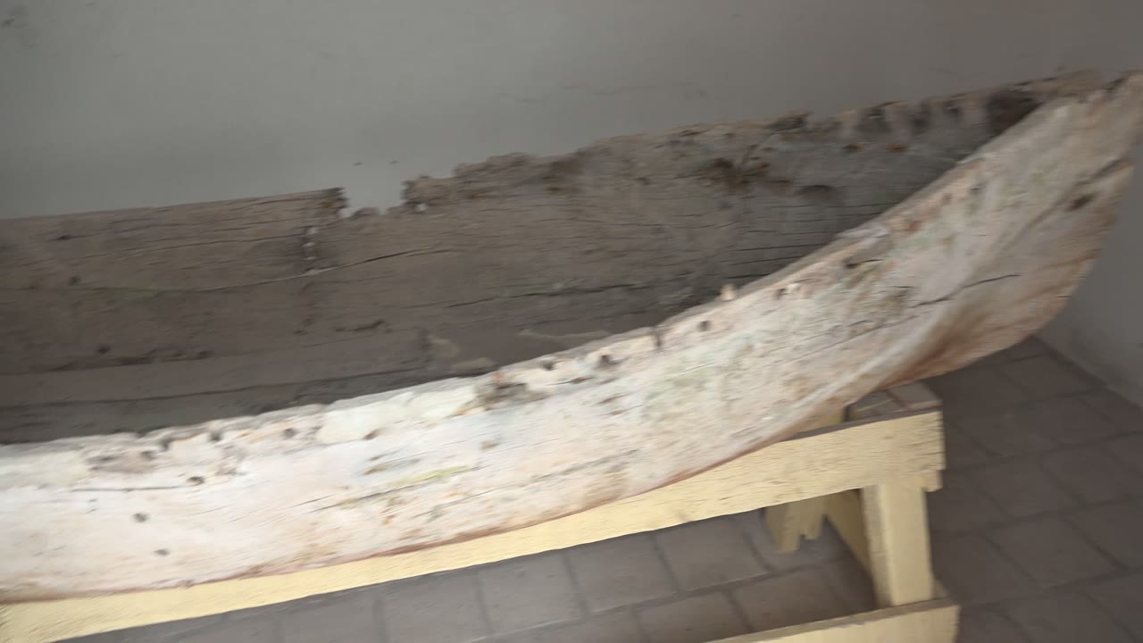 The camera view reaches the curved end of the weathered wooden boat in Mulege, with rough planks, a simple support frame, floor tiles, and no visible people.