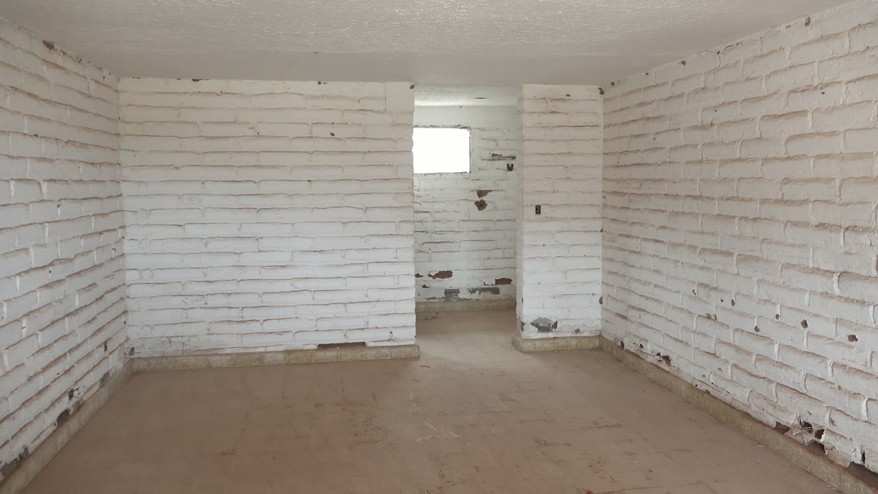Start of the Mulege interior clip showing an empty white brick room, concrete floor, dirty wall patches near the rear, a small bright window, and no visible people.