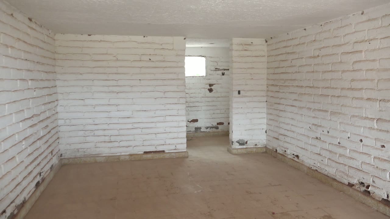 At 0:02, the Mulege room remains empty, with white painted brick on three visible walls, dark scuffs near the lower rear wall, a concrete floor, and no visible people.