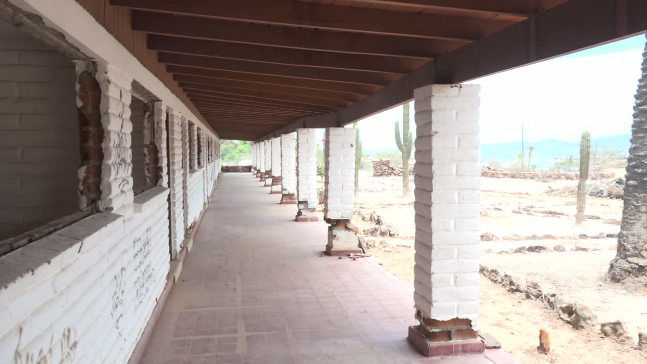 At 0:00 in Mulege, Baja California Sur, a shaded tiled walkway with white brick columns faces a bright desert yard with cacti, a palm tree, distant hills, and no visible people.