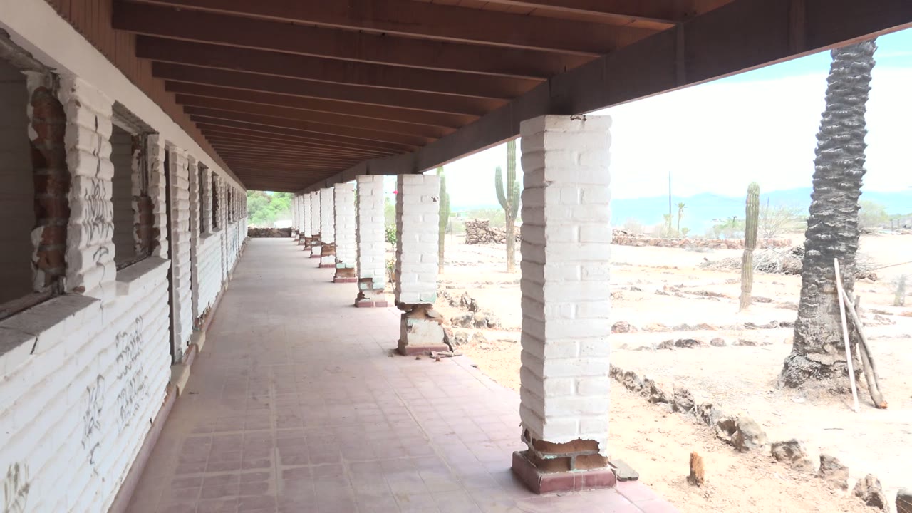 At 0:02 in Mulege, Baja California Sur, repeating white columns and a dark roof frame the shaded walkway while the overexposed desert ground, cactus, palm trunk, and no visible people fill the side view.