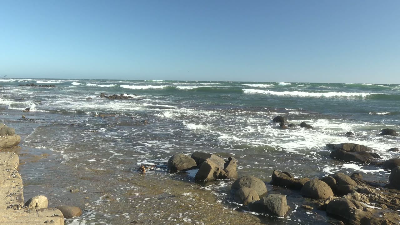 At La Bocana, Baja California Sur, white foam moves through shallow water around dark foreground rocks with distant surf bands and no visible people.