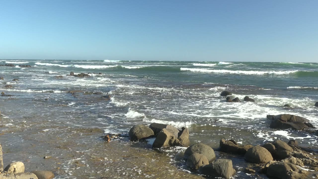 Foamy surf breaks around scattered rocks along a low shoreline.