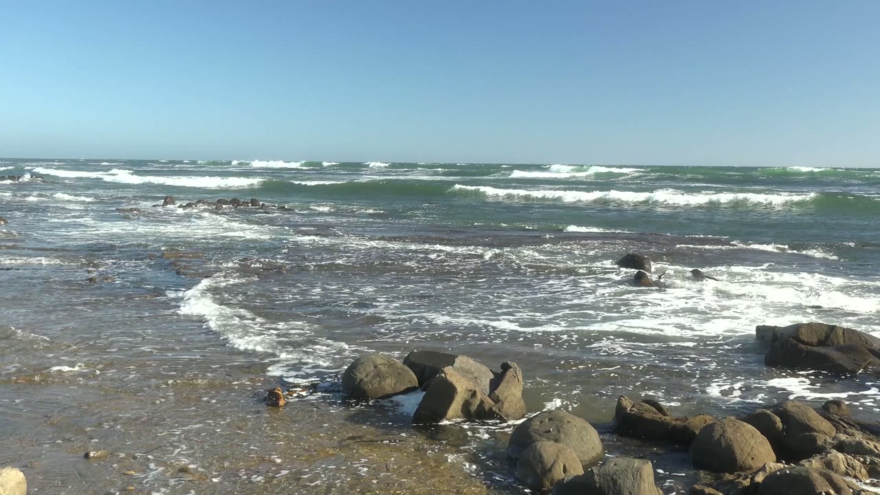 At La Bocana, Baja California Sur, broad whitewater spreads through the rocky shallows while the horizon remains clear under a blue sky, with no visible people.