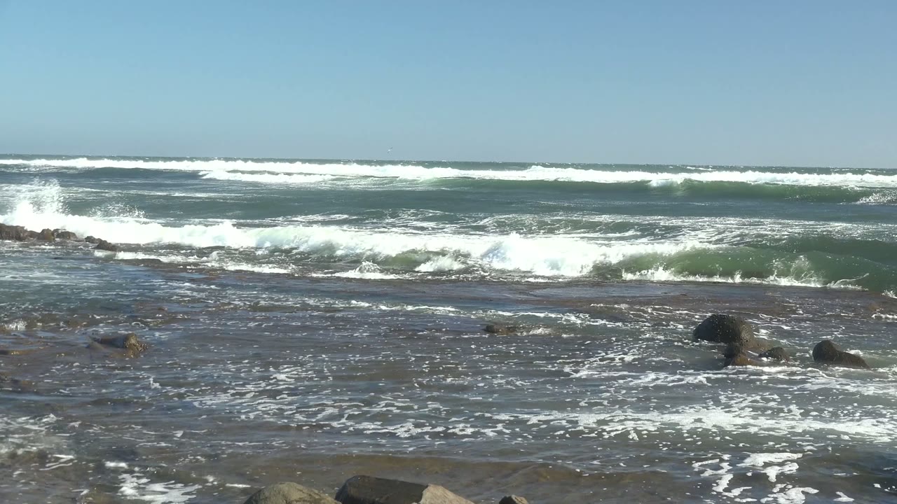 Wide view of blue-green surf and white foam crossing dark shoreline rocks in La Bocana, Baja California Sur, with no visible people.