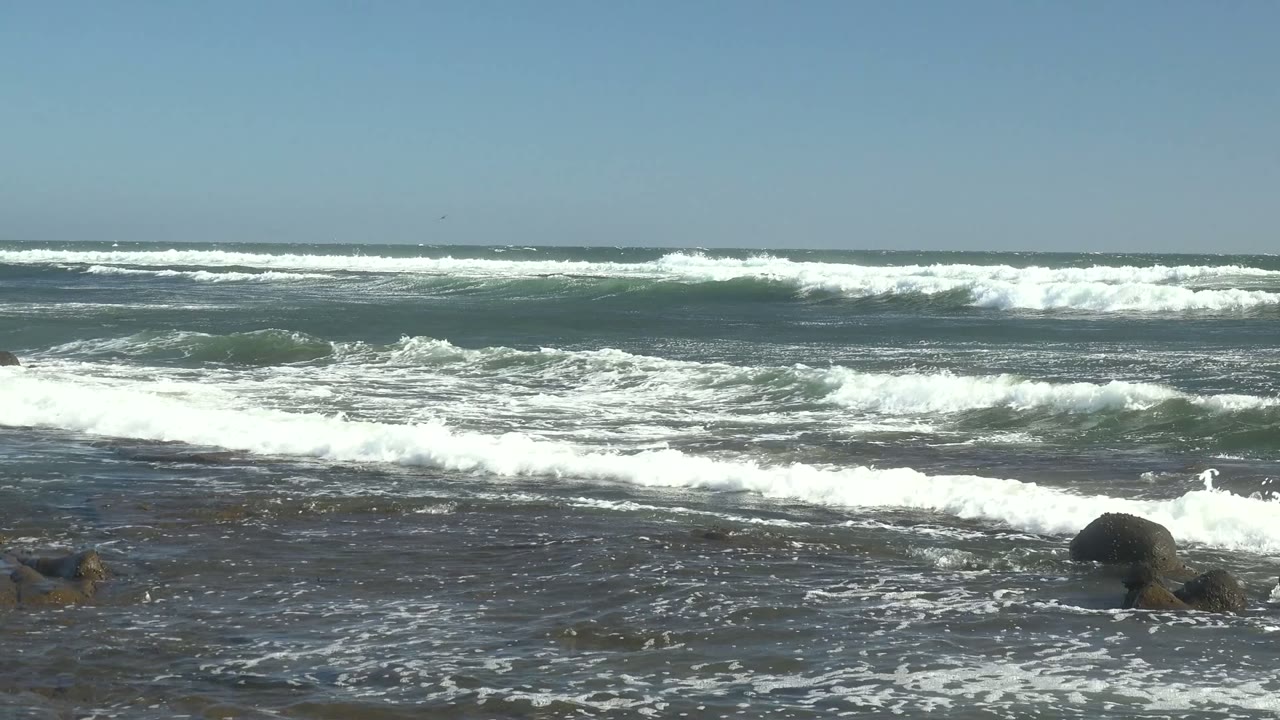 A low wide coastal view shows scattered dark rocks in shallow water as white surf breaks across the La Bocana shoreline, with no visible people.