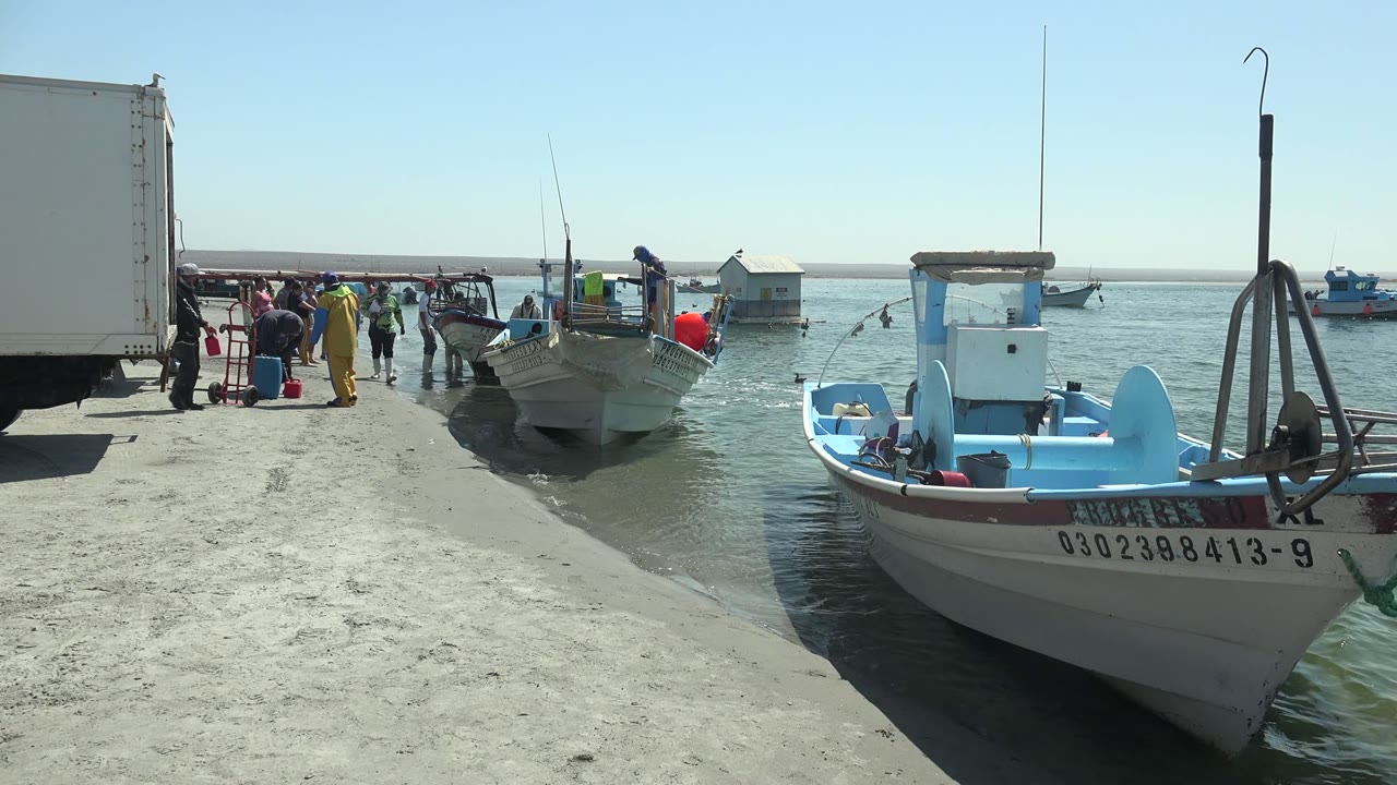 At La Bocana, Baja California Sur, the foreground boat fills the right side of frame as people remain visible near the docked fishing boat and white truck.