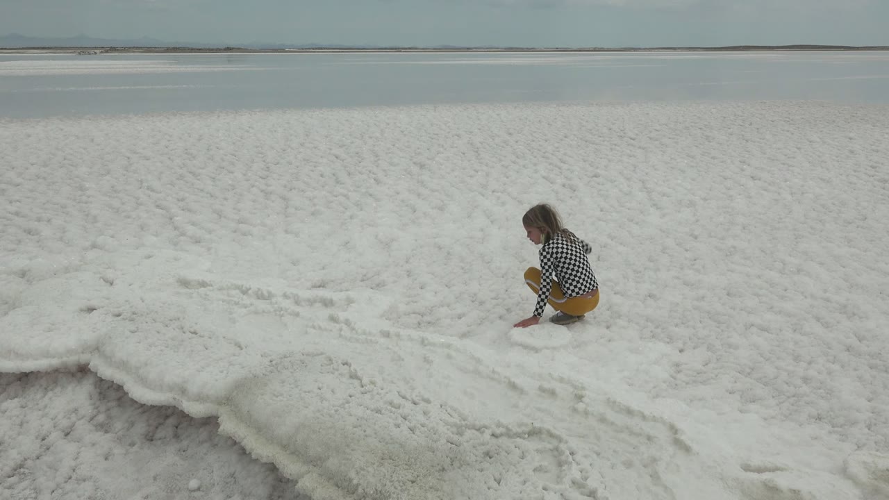 A person in yellow pants crouches on the crusted white salt flat at Guerrero Negro, Mexico, with calm water and a cloudy sky behind them.
