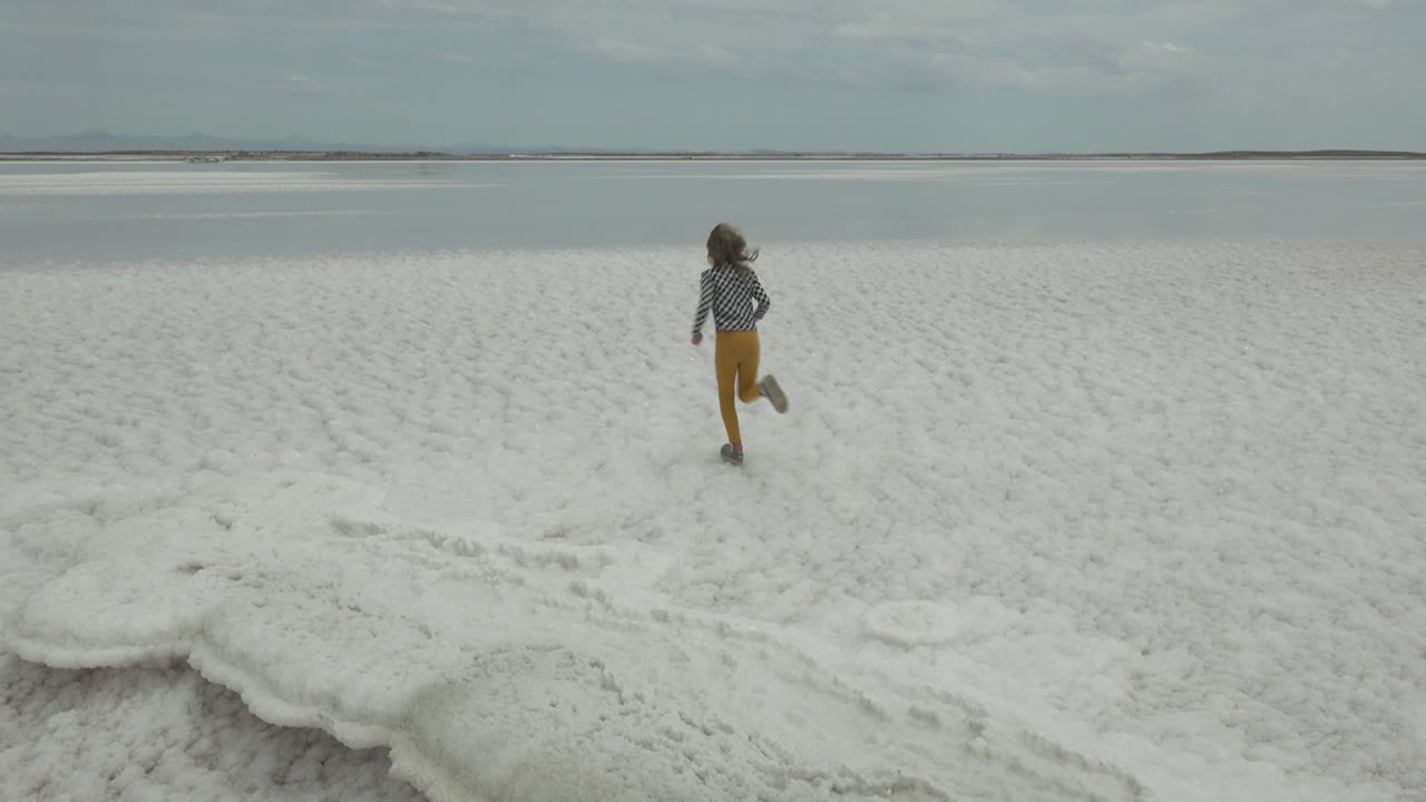 A small figure in a checkered shirt and yellow pants runs farther across the white salt flat at Guerrero Negro, Mexico, toward the distant water and sky.